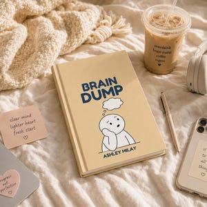 May include: A beige hardcover book titled Brain Dump by Ashley Milay with a cartoon character illustration, a pen, a smartphone with a sticker, a coffee cup with text, headphones, and two heart-shaped sticky notes with inspirational text.