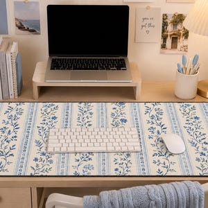 May include: A rectangular desk mat featuring a repeating pattern of vertical blue floral vines and thin striped lines on a cream background. The mat is positioned on a wooden desk surface beneath a white computer keyboard and a white computer mouse.