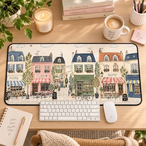 May include: A rectangular desk mat featuring a detailed illustration of a European street scene with storefronts labeled Fleurs, Café, Librairie, Bistro, and Boulanger. The design includes pastel-colored buildings, small trees, bicycles, and outdoor seating areas, all set against a light cream background with a black border.