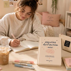 Puede incluir: Un libro de tapa dura beige titulado Executive Dysfunction Notes con una ilustraci&oacute;n de un cerebro, colocado sobre un escritorio junto a un port&aacute;til, un vaso de pl&aacute;stico con caf&eacute; helado, un estuche de lona y varios rotuladores pastel.