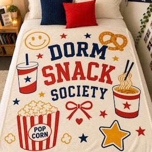 以下が含まれることがあります： 「DORM SNACK SOCIETY」という文字が青と赤の太字でプリントされたクリーム色のブランケット。スマイリーフェイス、プレッツェル、ドリンクカップ、箸付きの麺、ポップコーンのカラフルなグラフィックがデザインされています。
