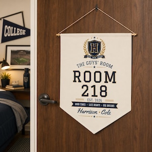 May include: A cream-colored fabric wall banner hanging from a wooden dowel with a string. It features a blue and gold crest with the letter H, the text THE GUYS' ROOM, ROOM 218, EST. 2026, GOOD TIMES, LATE NIGHTS, BIG DREAMS, and the names Harrison and Cole.