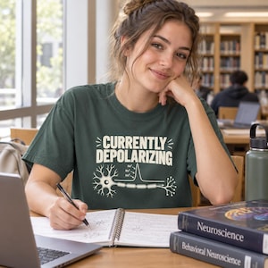 May include: A dark green short-sleeve t-shirt featuring the white text "CURRENTLY DEPOLARIZING" above a graphic illustration of a neuron and an action potential graph with sodium ion labels, printed on the front of the garment.