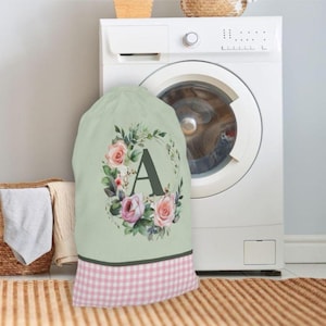 May include: A light green laundry bag with a floral wreath design and the letter "A". The wreath features pink roses and green leaves. The bottom of the bag has a pink and white gingham pattern. A washing machine is in the background.