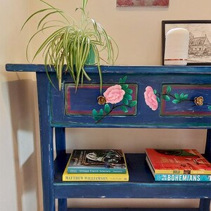 May include: A distressed blue console table with two drawers, decorated with pink floral designs and gold-coloured knobs. A green potted plant sits on top, with books on the lower shelf. The table is against a wall with a window.