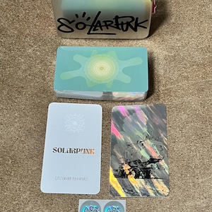May include: A silver metal tin with a pink elastic band and the text "SOLARPUNK" in black, a deck of cards with a green sun graphic, a white "SOLARPUNK CITIZENSHIP PASSPORT" card, a holographic card with a vehicle illustration, and two small holographic stickers.