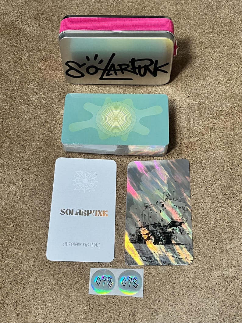 May include: A silver metal tin with a pink elastic band and the text "SOLARPUNK" in black, a deck of cards with a green sun graphic, a white "SOLARPUNK CITIZENSHIP PASSPORT" card, a holographic card with a vehicle illustration, and two small holographic stickers.