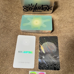 May include: A silver metal tin with the black text SOLARPUNK, a deck of cards with a green sun graphic, a white card labeled SOLARPUNK CITIZENSHIP PASSPORT, a card with a dark cityscape and sun, and two small circular holographic stickers with black symbols.