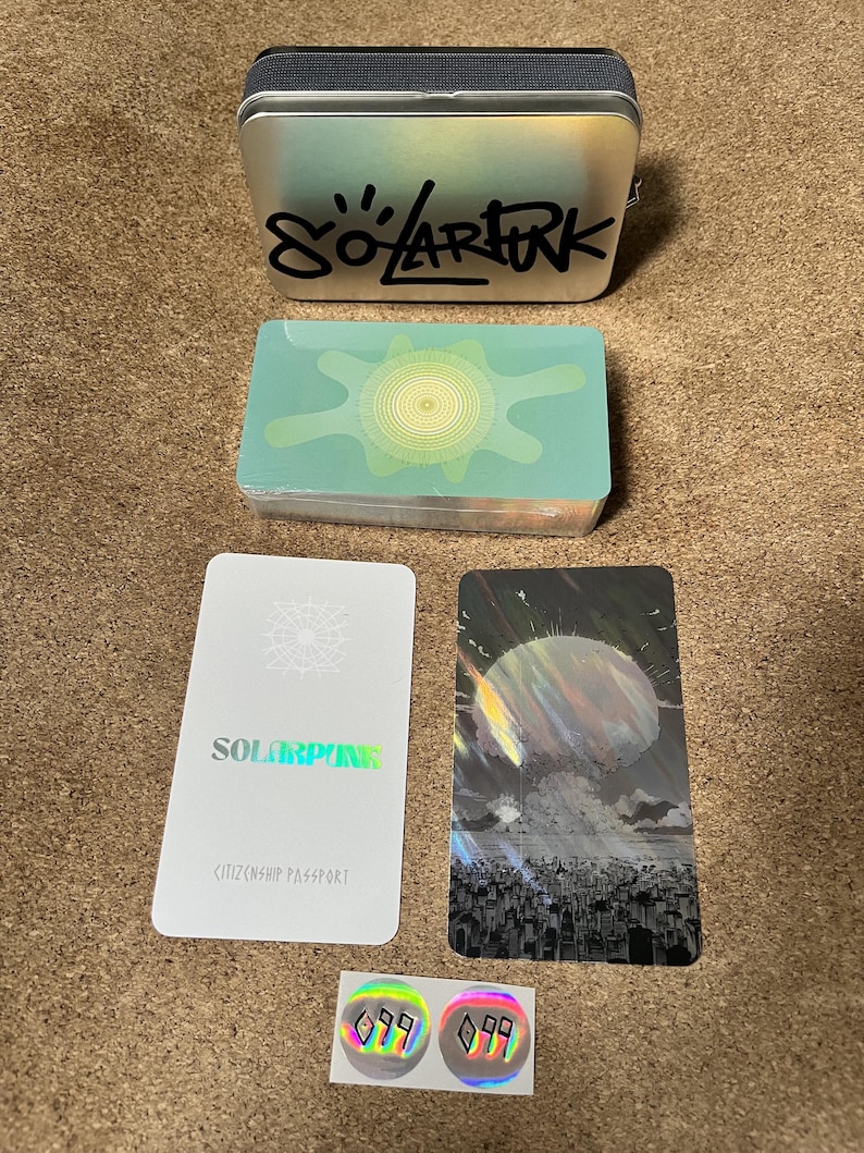 May include: A silver metal tin with the black text SOLARPUNK, a deck of cards with a green sun graphic, a white card labeled SOLARPUNK CITIZENSHIP PASSPORT, a card with a dark cityscape and sun, and two small circular holographic stickers with black symbols.