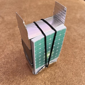 May include: A rectangular deck of cards featuring a green solar panel grid pattern on the front, secured with a black rubber band to a silver metallic structure made of foil-covered cardboard with two upright flaps extending from the top.