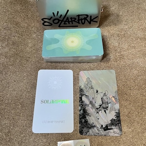 May include: A silver metal tin with an orange lid and the text SOLARFUN, a deck of cards with a light blue abstract design, a white card labeled SOLARFUN CITIZENSHIP PASSPORT, a holographic card featuring a figure in a landscape, and two circular 100 stickers.