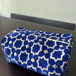 Op de afbeelding: Een rechthoekige tas met rits van diepblauw fluweelachtig materiaal, voorzien van een geometrisch patroon van zilverwitte achtpuntige sterren en kleine gouden ruitvormige accenten. De tas heeft een gestructureerde vorm met een ritssluiting.