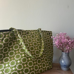 Può includere: Una grande borsa tote verde con un motivo geometrico bianco e oro. La borsa ha manici abbinati e poggia su una superficie di legno. Un piccolo vaso blu con fiori rosa è a destra.