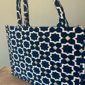 May include: A navy blue tote bag with a geometric star pattern in white and gold. The bag has matching navy blue handles with a gold trim. The bag is made of a textured fabric.