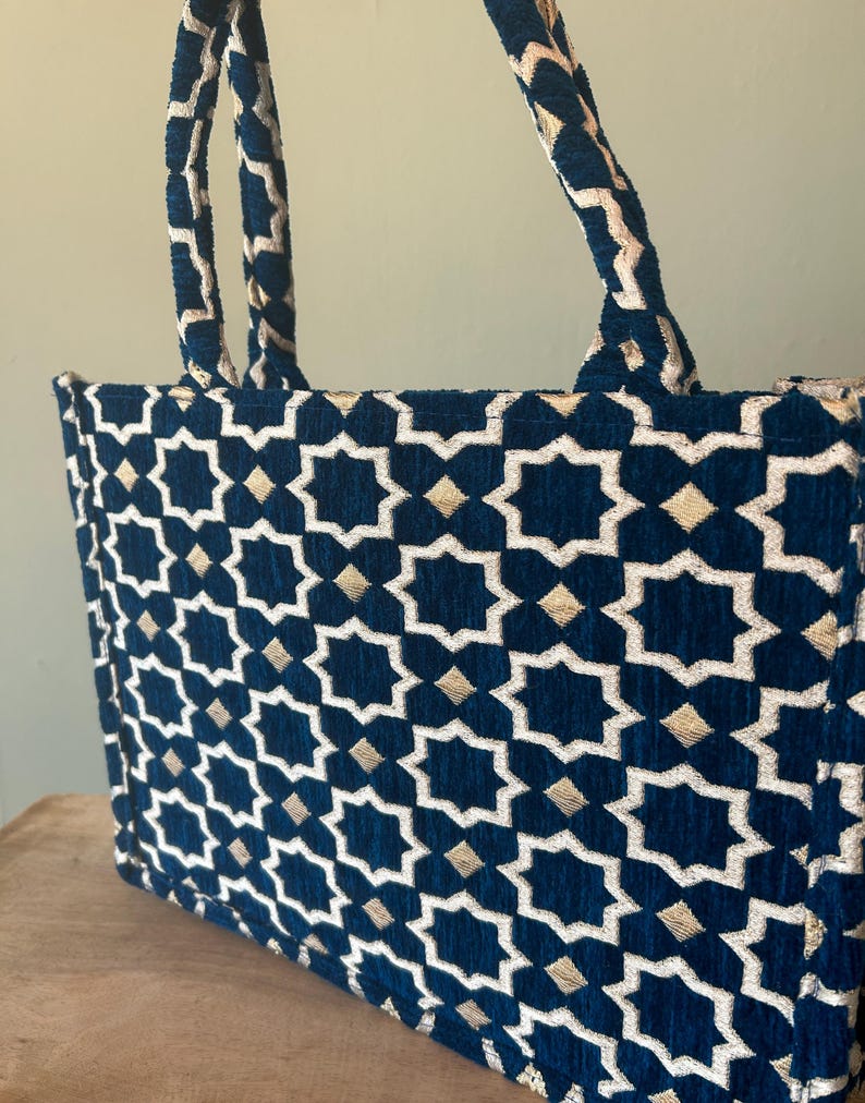 May include: A navy blue tote bag with a geometric star pattern in white and gold. The bag has matching navy blue handles with a gold trim. The bag is made of a textured fabric.