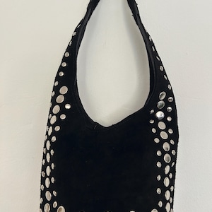 Op de afbeelding: Zwarte suède schoudertas versierd met talrijke zilverkleurige, cirkelvormige studs. De tas heeft een gebogen, halvemaanvormige vorm met een enkele schouderriem. De studs zijn in een verspreid patroon over de voorkant en zijkanten geplaatst.