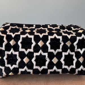 Op de afbeelding: Een rechthoekige tas met ritssluiting van zwarte, fluweelachtige stof met een herhalend geometrisch patroon van witte achtpuntige sterren en kleine beige ruitvormige accenten. De tas is voorzien van een handvat aan de zijkant en een rits.