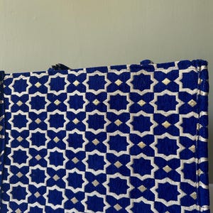 May include: A rectangular tote bag with a geometric pattern. The bag features a deep blue background with a repeating pattern of white and gold star-like shapes. The bag has a structured design and appears to be made of a textured fabric.