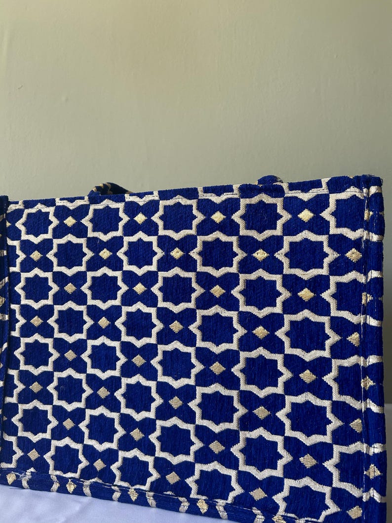 May include: A rectangular tote bag with a geometric pattern. The bag features a deep blue background with a repeating pattern of white and gold star-like shapes. The bag has a structured design and appears to be made of a textured fabric.