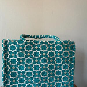 May include: A teal and gold patterned handbag with a matching handle. The bag features a repeating geometric star design. The handle is a twisted rope style. The bag is rectangular and appears to be made of a textured fabric.