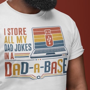 Op de afbeelding: Wit t-shirt met een retro graphic. Het ontwerp toont een laptop met een USB-symbool en de tekst "I STORE ALL MY DAD JOKES IN A DAD-A-BASE". Het shirt heeft een ronde hals en korte mouwen.
