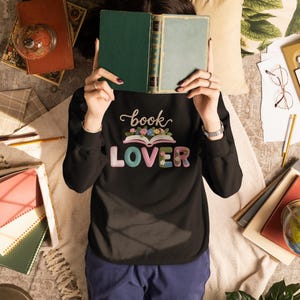 Op de afbeelding: Een zwart sweatshirt met de woorden "book LOVER" in kleurrijke letters en een bloemenontwerp. Een persoon houdt een groen en blauw boek vast. Boeken, een wereldbol en andere items zijn verspreid.