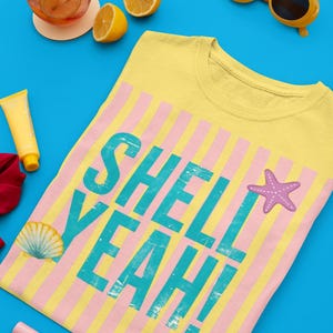May include: Yellow t-shirt with pink and yellow stripes and the words "SHELL YEAH!" in turquoise. A seashell and starfish graphic are also on the shirt. The shirt is on a blue surface, with sunglasses, a drink, and lemons.