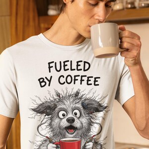 Puede incluir: Camiseta blanca con el texto "FUELED BY COFFEE" y una ilustración de dibujos animados de un perro con cables eléctricos y una taza de café roja. La persona de la imagen está bebiendo de una taza de café beige.