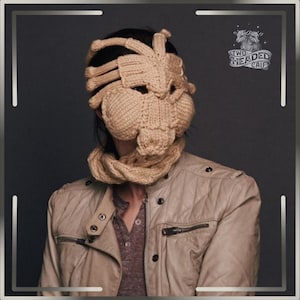 May include: A beige knitted mask and scarf set, obscuring the face. The mask features a rib-like design, with rounded sections covering the cheeks and chin. The person is wearing a tan leather jacket with a button-down shirt underneath.
