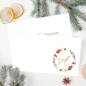 May include: Two white rectangular cards. The top card features the letter A in gold script, centered within a circular wreath of watercolor autumn leaves, berries, and sprigs. The partially visible bottom card displays the text THANKSGIVING.