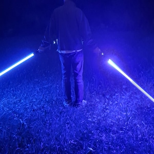Könnte beinhalten: Eine Person hält zwei leuchtende blaue Lichtschwerter in einem grasbewachsenen Bereich bei Nacht. Die Lichtschwerter strahlen hellblaues Licht aus, das mit der dunklen Umgebung kontrastiert. Die Szene wird vom Licht der Lichtschwerter erhellt.