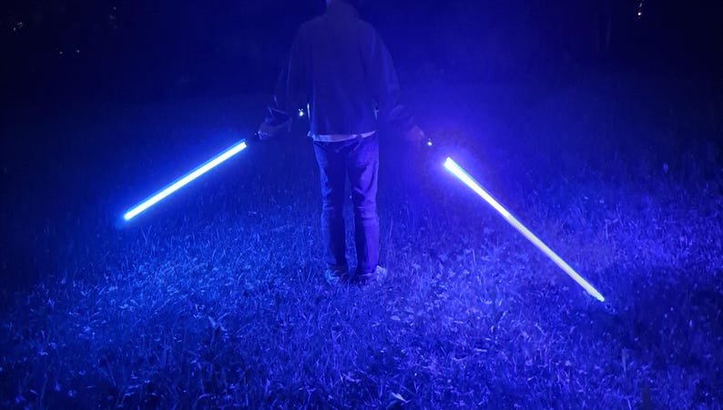 May include: A person stands holding two blue lightsabers in a grassy area at night. The lightsabers glow brightly, casting a blue light against the dark background. The scene is lit by the lightsabers' illumination.