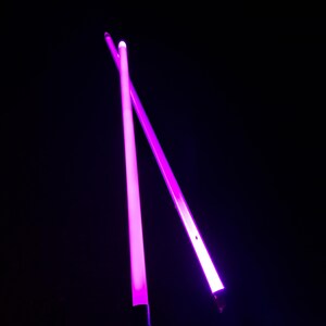May include: Two neon pink light sabers, lit against a black backdrop. The sabers are crossed, with a bright glow from the blades. The image is dark, with the sabers as the main focus, creating a striking contrast.
