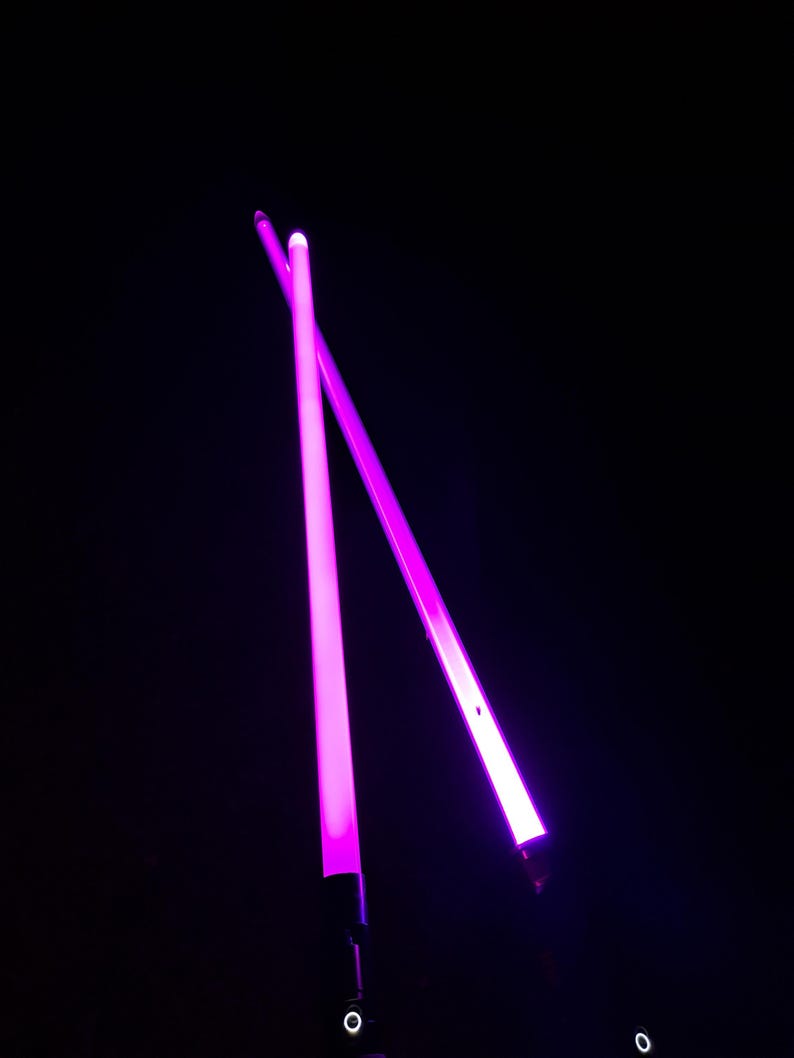 May include: Two neon pink light sabers, lit against a black backdrop. The sabers are crossed, with a bright glow from the blades. The image is dark, with the sabers as the main focus, creating a striking contrast.
