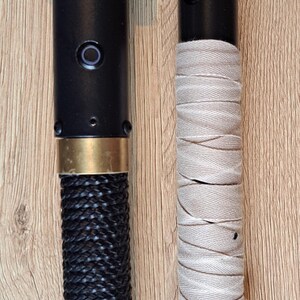 May include: Two custom lightsabers. One has a black braided handle and gold details, the other a wrapped beige grip and a white blade. Both have black cylindrical bodies, displayed on a wooden surface.