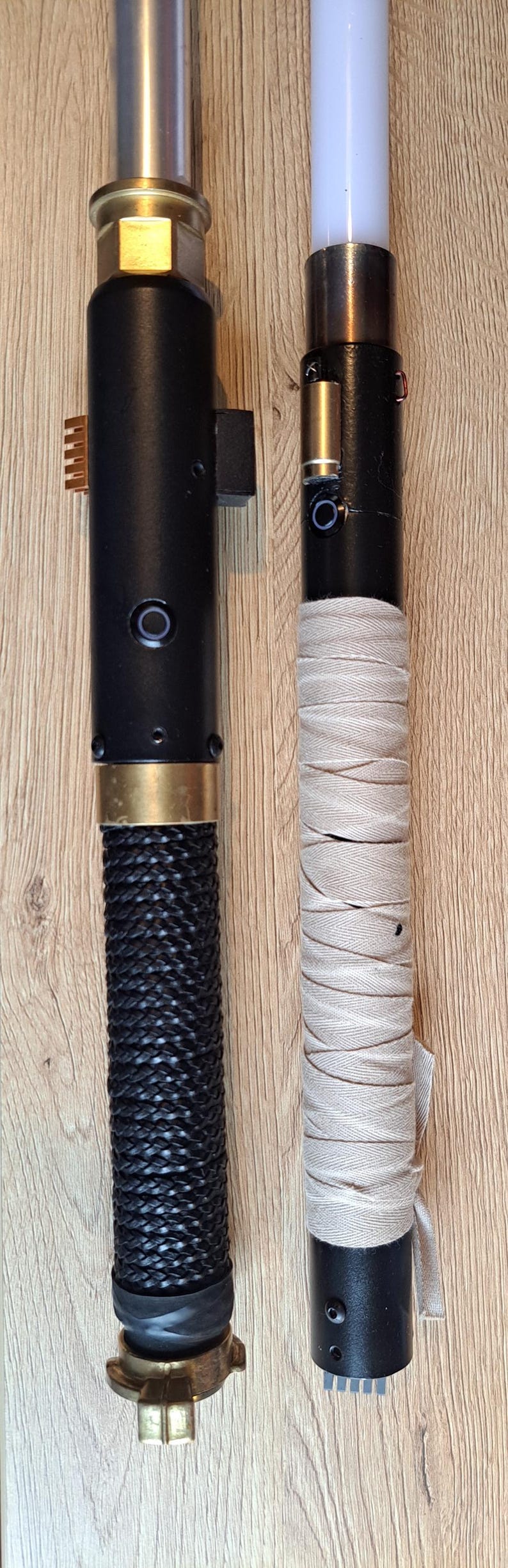 May include: Two custom lightsabers. One has a black braided handle and gold details, the other a wrapped beige grip and a white blade. Both have black cylindrical bodies, displayed on a wooden surface.
