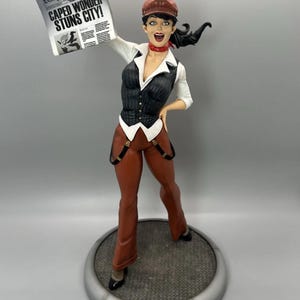May include: A figurine of a reporter wearing a brown cap, white shirt, black pinstriped vest, red neckerchief, and brown trousers with suspenders. The figure holds a Daily Planet newspaper with the headline CAPED WONDER STUNS CITY! above its head.