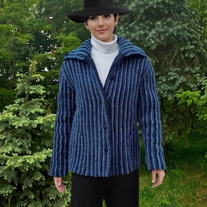 Può includere: Giacca blu a coste con colletto a revers e chiusura con bottoni. La giacca è indossata sopra un dolcevita bianco e abbinata a pantaloni neri. Un cappello nero completa l'outfit. Lo sfondo presenta un fogliame verde.