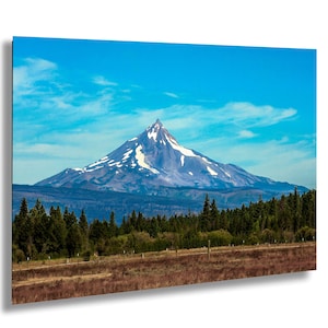 Op de afbeelding: Een landschapsfoto van een besneeuwde bergtop tegen een helderblauwe lucht met verspreide wolken. De voorgrond toont een bos en een veld, wat een natuurlijke scène creëert.