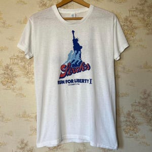 May include: White t-shirt with a graphic of the Statue of Liberty and runners in blue and red. The text "Stroh's Run For Liberty I October 13, 1984" is printed below the graphic. The shirt is displayed on a wooden hanger.