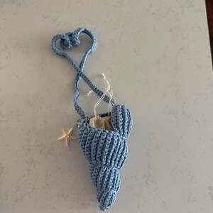 May include: A light blue crochet seashell-shaped bag with a long strap and a small starfish charm. The bag has a drawstring closure and a small pouch inside. The strap forms a heart shape at the top.