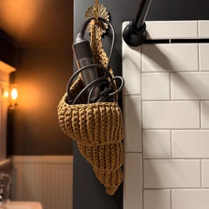May include: A tan, crochet seashell-shaped storage basket hanging on a dark wall. A silver hair dryer is stored inside the basket. The basket is suspended from a decorative gold hook.