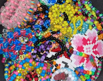 Può includere: Una collezione di braccialetti e accessori colorati con perline. L'assortimento comprende braccialetti in vari colori come rosa, giallo, blu e nero. Alcuni braccialetti presentano motivi e ciondoli. L'immagine mostra una varietà di gioielli fatti a mano.