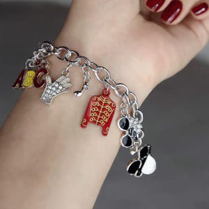 Op de afbeelding: Zilveren bedelarmband met diverse bedels, waaronder een rode jas, een muzieknoot, een met juwelen bezette hand en een zwart-wit ontwerp. De armband is gemaakt van zilveren schakels.