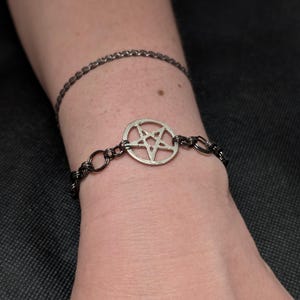 May include: A silver-colored pentagram charm bracelet with a dark chain. A second, thinner chain bracelet is visible above it. The bracelets are worn on a wrist, with a dark background.