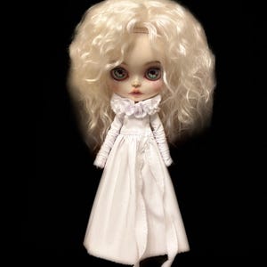 May include: A doll with a pale complexion, large green eyes, and a white dress. The doll has a large head of curly, light-coloured hair. The dress features a ruffled collar and long sleeves. The doll is set against a black background.