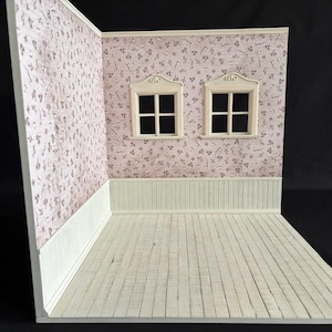 Könnte beinhalten: Ein Miniatur-Puppenhauszimmer mit hellrosa floraler Tapete und weißer Verkleidung. Zwei kleine Fenster sind an der Rückwand zentriert. Der Boden besteht aus weißen Holzplanken. Der Raum ist leer.