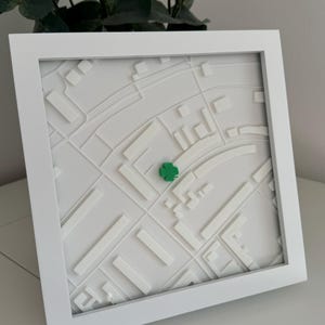 Op de afbeelding: Een wit omkaderd 3D-kaartkunstwerk. De kaart is een reliëf van een stadsplattegrond, met witte verhoogde structuren en wegen. Een klein groen klavertje is op de kaart geplaatst. De lijst is vierkant en wit.