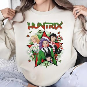 May include: Cream-colored sweatshirt featuring a graphic of three women in front of a red star. The word "HUNTRIX" is above the graphic in red and green. The women are wearing holiday-themed clothing and holding gifts.