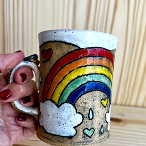 May include: A ceramic mug with a rainbow design. The mug features a white rim and handle, with a colorful rainbow, clouds, hearts, and raindrops painted on the exterior. The mug is held by a person.